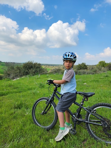 A young child wearing a blue helmet and casual outfit is standing with their bicycle on a grassy field. The background is a scenic landscape with a clear blue sky filled with fluffy clouds and green, bushy vegetation.