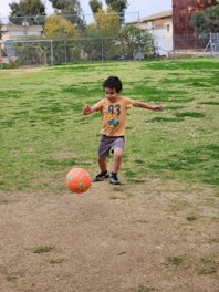 A young child wearing a yellow t-shirt and shorts is playing soccer on a grassy field. The child is kicking a brightly colored orange soccer ball. There are trees and buildings in the background, with a chain-link fence surrounding the area.
