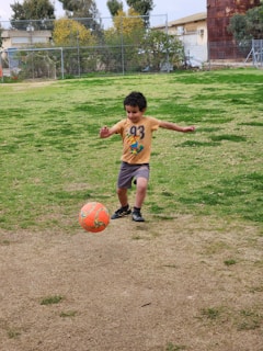 A young child wearing a yellow t-shirt and shorts is playing soccer on a grassy field. The child is kicking a brightly colored orange soccer ball. There are trees and buildings in the background, with a chain-link fence surrounding the area.