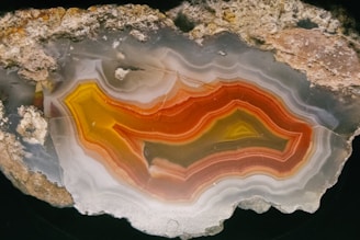 A striking cross-section of a geode with intricate bands of orange, red, yellow, and gray. The layers are detailed and showcase patterns resembling contours or waves through the stone, contrasting against the rough outer rocky surface.