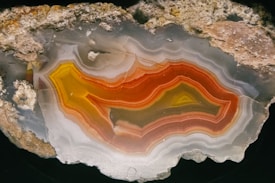 A striking cross-section of a geode with intricate bands of orange, red, yellow, and gray. The layers are detailed and showcase patterns resembling contours or waves through the stone, contrasting against the rough outer rocky surface.