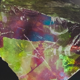 A vibrant mineral or crystal with a rough and uneven texture, showcasing a mixture of colors including green, blue, red, and yellow. The surface has a glassy appearance with natural fractures and imperfections, reflecting light in various directions.