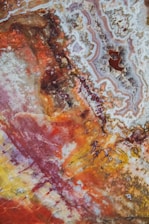 An abstract image resembling colorful mineral or rock formations. There are swirling patterns and mixed textures predominantly in red, yellow, orange, and brown shades. Intricate details and organic shapes are visible throughout the image, creating a visually complex and vibrant scene.