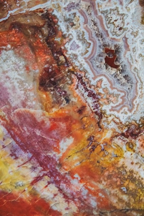 An abstract image resembling colorful mineral or rock formations. There are swirling patterns and mixed textures predominantly in red, yellow, orange, and brown shades. Intricate details and organic shapes are visible throughout the image, creating a visually complex and vibrant scene.