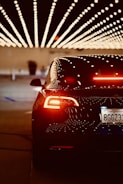A sleek, black car with illuminated taillights is positioned under a ceiling decorated with rows of bright, dotted lights, creating a visually striking pattern. The license plate indicates a California registration.