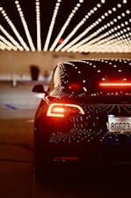 A sleek, black car with illuminated taillights is positioned under a ceiling decorated with rows of bright, dotted lights, creating a visually striking pattern. The license plate indicates a California registration.