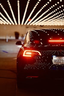 A sleek, black car with illuminated taillights is positioned under a ceiling decorated with rows of bright, dotted lights, creating a visually striking pattern. The license plate indicates a California registration.