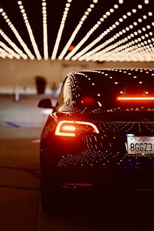 A sleek, black car with illuminated taillights is positioned under a ceiling decorated with rows of bright, dotted lights, creating a visually striking pattern. The license plate indicates a California registration.