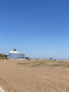 A sandy beach stretches out under a clear blue sky, with sparse grass patches in the foreground. A large, multi-story building with a distinctive dome-shaped roof stands near the beach, featuring numerous balconies. In the distance, a few small structures and a solitary bench are visible.