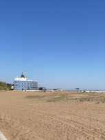 A sandy beach stretches out under a clear blue sky, with sparse grass patches in the foreground. A large, multi-story building with a distinctive dome-shaped roof stands near the beach, featuring numerous balconies. In the distance, a few small structures and a solitary bench are visible.