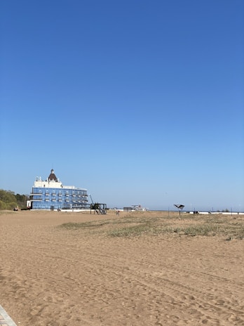A sandy beach stretches out under a clear blue sky, with sparse grass patches in the foreground. A large, multi-story building with a distinctive dome-shaped roof stands near the beach, featuring numerous balconies. In the distance, a few small structures and a solitary bench are visible.