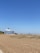 A sandy beach stretches out under a clear blue sky, with sparse grass patches in the foreground. A large, multi-story building with a distinctive dome-shaped roof stands near the beach, featuring numerous balconies. In the distance, a few small structures and a solitary bench are visible.