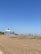 A sandy beach stretches out under a clear blue sky, with sparse grass patches in the foreground. A large, multi-story building with a distinctive dome-shaped roof stands near the beach, featuring numerous balconies. In the distance, a few small structures and a solitary bench are visible.