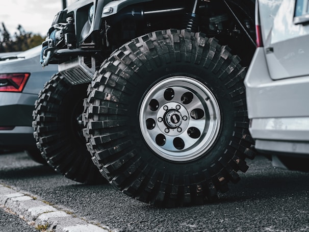Two vehicles are parked side by side on an asphalt road. The focus is on the large, rugged off-road tires equipped on one of the vehicles, which appears to be customized for off-road or rough terrain driving. The other vehicle has standard road tires. A curb is visible in the foreground.