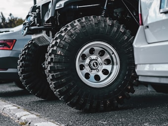 Two vehicles are parked side by side on an asphalt road. The focus is on the large, rugged off-road tires equipped on one of the vehicles, which appears to be customized for off-road or rough terrain driving. The other vehicle has standard road tires. A curb is visible in the foreground.