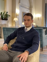 A man is seated in an elegant, well-furnished room featuring a classical style. He is wearing a navy blue double-breasted blazer, white shirt, and a patterned tie. The room has sophisticated décor with ornate molding, lamps, and plants. An upholstered chair and soft lighting create a cozy, luxurious atmosphere.