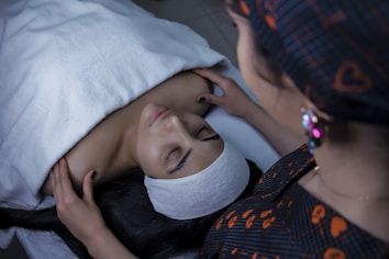 A person is lying down on a treatment bed covered with a white towel. Another individual is standing beside, placing their hands on the person's shoulders as if about to perform a massage or therapy. The scene appears to be in a spa or similar wellness setting.