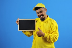A person wearing a bright yellow shirt and cap holds a small blank chalkboard. The individual is smiling and giving a thumbs-up gesture with their other hand against a plain blue background.