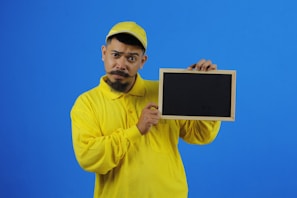 A person in a yellow cap and shirt holds a small chalkboard against a blue background. The individual's facial expression conveys a sense of curiosity or questioning.