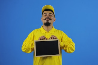 A person wearing a yellow shirt and cap holds a small blank chalkboard against a plain blue background. The individual has a styled mustache and beard, and their facial expression appears confident.