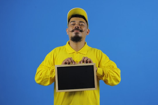 A person wearing a yellow shirt and cap holds a small blank chalkboard against a plain blue background. The individual has a styled mustache and beard, and their facial expression appears confident.