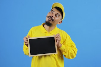 A man with a mustache is wearing a yellow shirt and cap. He is holding a blank blackboard with both hands and tilting his head slightly to the side. The background is a solid blue color.