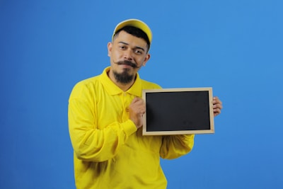 A person wearing a bright yellow shirt and cap is holding a rectangular blank chalkboard against a solid blue background. The individual has a distinctive facial hair style and a friendly expression.