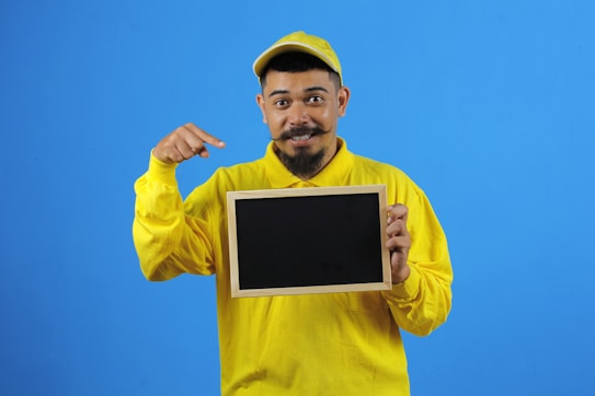A person wearing a yellow cap and shirt is holding a small blackboard and pointing at it with a smile. The background is a solid blue color.