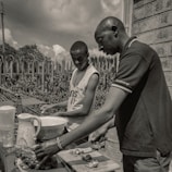 Two individuals are outdoors engaged in food preparation. They are focused on cutting and arranging pieces of food on a table that includes a bowl, pitcher, and chopping board. The setting is casual with a wire fence and foliage in the background under a partly cloudy sky.