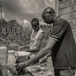 Two individuals are outdoors engaged in food preparation. They are focused on cutting and arranging pieces of food on a table that includes a bowl, pitcher, and chopping board. The setting is casual with a wire fence and foliage in the background under a partly cloudy sky.