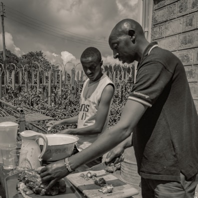 Two individuals are outdoors engaged in food preparation. They are focused on cutting and arranging pieces of food on a table that includes a bowl, pitcher, and chopping board. The setting is casual with a wire fence and foliage in the background under a partly cloudy sky.