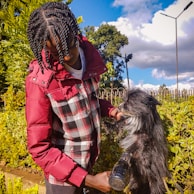 A person in a red and plaid jacket is grooming a small black and white dog. They appear to be using a hairdryer on the dog's fur. The scene is outdoors with greenery and a clear blue sky with some clouds visible in the background.