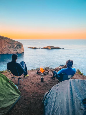 Outdoor cooking setup with campfire and rugged chairs on a coastal cliff.