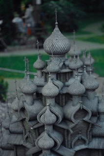 A detailed architectural model featuring numerous onion-shaped domes, reminiscent of Russian or Eastern European style buildings. The structure is crafted with intricate patterns, giving an impression of a wooden texture. Surrounding the model is a green, blurred background, suggesting an outdoor setting possibly in a park.