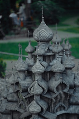 A detailed architectural model featuring numerous onion-shaped domes, reminiscent of Russian or Eastern European style buildings. The structure is crafted with intricate patterns, giving an impression of a wooden texture. Surrounding the model is a green, blurred background, suggesting an outdoor setting possibly in a park.