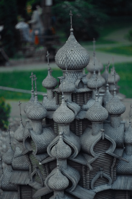A detailed architectural model featuring numerous onion-shaped domes, reminiscent of Russian or Eastern European style buildings. The structure is crafted with intricate patterns, giving an impression of a wooden texture. Surrounding the model is a green, blurred background, suggesting an outdoor setting possibly in a park.