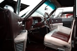 The interior of a vintage car with an open door. The seats are upholstered in a combination of white and burgundy leather. The dashboard and steering wheel feature classic design elements, with a visible gear shift and analog gauges. The car's exterior through the windows appears to be a classic design as well.