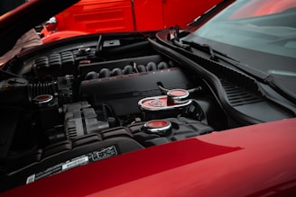 A close-up view of a car engine bay, emphasizing a sleek, black engine cover with the word 'Corvette' and the number '6.2L' prominently visible. The engine components are surrounded by glossy red body panels, hinting at a high-performance vehicle.