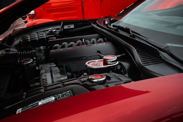 A close-up view of a car engine bay, emphasizing a sleek, black engine cover with the word 'Corvette' and the number '6.2L' prominently visible. The engine components are surrounded by glossy red body panels, hinting at a high-performance vehicle.