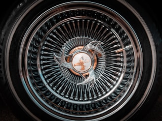 An intricately designed car wheel rim featuring a complex pattern of concentric circles and a central emblem. The metallic surface reflects light, creating a shiny, detailed appearance.