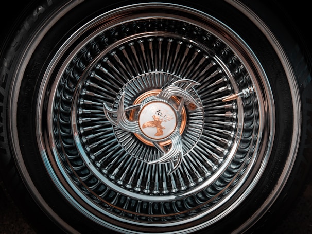 An intricately designed car wheel rim featuring a complex pattern of concentric circles and a central emblem. The metallic surface reflects light, creating a shiny, detailed appearance.