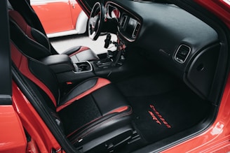 The interior of a car featuring black and red leather seats, a detailed dashboard with a steering wheel, a gear shift, and a center console with cup holders. The door trim and floor mat are also visible, with the mat displaying the words 'CHARGER SRT'. Another red car is partially visible through the window.