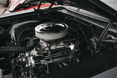 A close-up view of a car engine with various components such as hoses, a fan belt, and a shiny air filter. The engine appears well-maintained, showcasing metallic and rubber textures. The background includes a glimpse of a red car body.