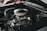 A close-up view of a car engine with various components such as hoses, a fan belt, and a shiny air filter. The engine appears well-maintained, showcasing metallic and rubber textures. The background includes a glimpse of a red car body.