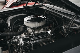A close-up view of a car engine with various components such as hoses, a fan belt, and a shiny air filter. The engine appears well-maintained, showcasing metallic and rubber textures. The background includes a glimpse of a red car body.