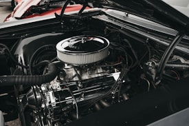 A close-up view of a car engine with various components such as hoses, a fan belt, and a shiny air filter. The engine appears well-maintained, showcasing metallic and rubber textures. The background includes a glimpse of a red car body.