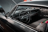 A vintage car interior with a focus on the steering wheel, dashboard, and front seats. The dashboard features classic dials and buttons, and the upholstery is well-maintained, showcasing a retro design. A beaded necklace or string hangs from the rearview mirror, adding a touch of personalization.