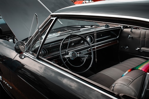 A vintage car interior with a focus on the steering wheel, dashboard, and front seats. The dashboard features classic dials and buttons, and the upholstery is well-maintained, showcasing a retro design. A beaded necklace or string hangs from the rearview mirror, adding a touch of personalization.