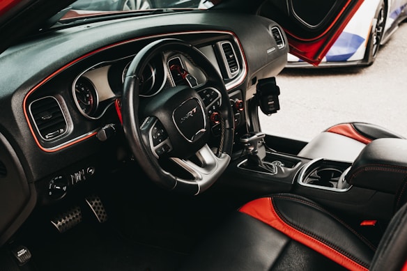 The interior of a car featuring a steering wheel with the SRT logo, black and red leather seats, and a dashboard equipped with round gauges and control knobs. The driver's side door is open, revealing parts of another vehicle outside.