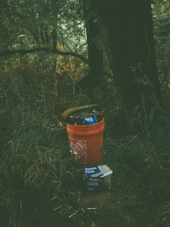 An orange bucket with a brand logo is placed on a patch of grass in a forested area. Inside the bucket are various tools or items, and next to it, there is a box labeled 'Fastenal.' The background features dense greenery and trees, with the lighting creating a shadowy and mysterious atmosphere.